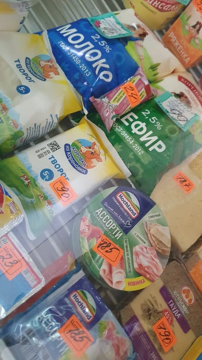 This is a close-up, overhead shot of a refrigerated display case containing various dairy products for sale, likely in a grocery store. The lighting is artificial and casts a slight glare on the plastic packaging. The items are arranged in a slightly disorganized manner, with some overlapping.
Prominent items include:
- Several packages of "Moloko" (milk), one blue and white with "2.5%" and "GOST 31450-2013" printed on it. Another package, yellow and white, is labeled "Tvorog" (cottage cheese) with "5%" and "Iz Korenovki" (from Korenovka) with a cow logo.
- A green package labeled "Kefir" (kefir) with "2.5%" and "GOST 31454-2012".
- A round, green package of "Assorti" (assortment) processed cheese by "Hochland", with an image of ham and cheese.
- A package of "Ryazhenka" (baked milk) in an orange wrapper.
- A package of "Gouda" cheese from "Yugovskoy" with "45%" and an orange price tag.
- Several smaller, pink-wrapped items.
Orange price tags are visible on many of the products, displaying prices like "90", "78", "23", "85", "190".
The background consists of horizontal metal shelving, typical of a refrigerated display. The overall impression is of a well-stocked but somewhat cluttered dairy section of a store. The image does not provide any specific visual cues to pinpoint its location within Ust-Labinsk, Russia, beyond the typical appearance of a Russian grocery store dairy aisle.