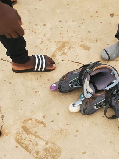 The image shows a close-up view of a person's feet and a pair of roller skates on a concrete surface. One foot is wearing a black slide sandal with three white stripes. The other foot is covered by a grey sock with a black band at the top. A pair of grey and black roller skates are lying on the ground, with one skate partially disassembled. The concrete surface appears wet in some areas and has several dark stains. The background is out of focus, but suggests an outdoor environment. This scene is located in Pankshin, Nigeria.