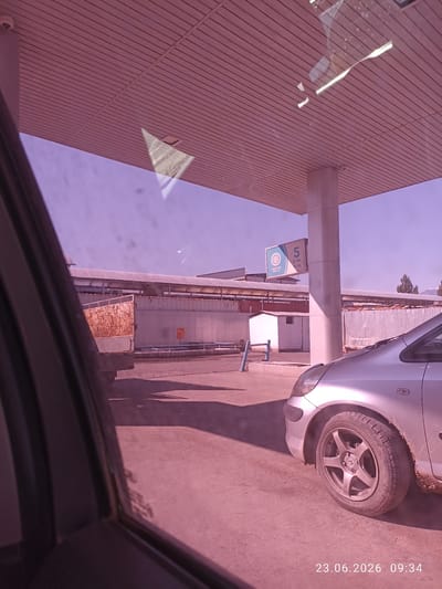 A silver sedan is parked at a gas station in Batken City, Kyrgyzstan. The car is in the foreground on the right side of the image, with its front tire and fender visible. The gas station has a covered canopy with a sign displaying the number "5" and fuel prices. In the background, there are industrial buildings and a truck. The sky is clear and the lighting suggests it is daytime. A date and time stamp in the bottom right corner reads "23.06.2026 09:34".