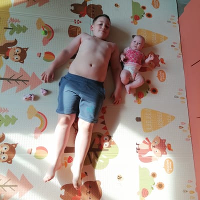 Two children are lying on their backs on a play mat. On the left is a boy, shirtless, with dark hair, wearing blue denim shorts. He is looking towards the camera with a slight smile. To his right is a baby, wearing a pink and white strawberry-patterned top and a diaper. The baby is looking towards the camera with wide eyes. The play mat has a colorful pattern of cartoon woodland animals, trees, and flowers. In the background, to the right of the children, a pink object, likely a wall or a piece of furniture, is visible. The lighting suggests it is daytime, with sunlight casting shadows across the play mat. The overall scene is casual and appears to be in a home environment, possibly a nursery or playroom. There is no visible text in the image.