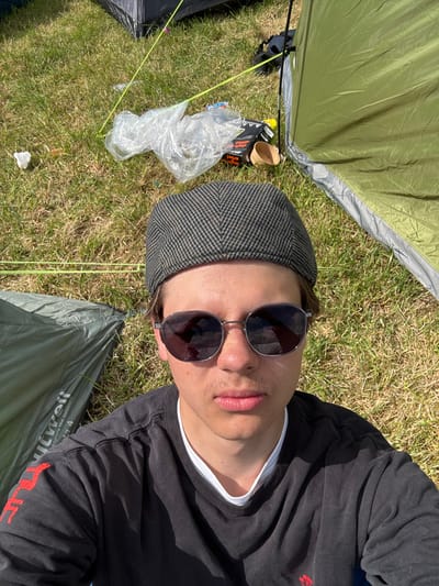 A person wearing sunglasses and a patterned cap lies on grass amidst camping equipment. The person is wearing a dark, long-sleeved shirt with a white undershirt visible at the collar. Tents, tent stakes, and some discarded items like a plastic bag and cardboard containers are scattered on the grass. The scene appears to be an outdoor camping setting under bright sunlight.