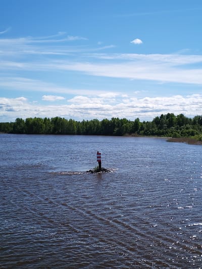 The image shows a body of water under a blue sky with scattered clouds. A forested shoreline is visible in the background. In the middle ground, a striped red and white post stands on a small rocky base in the water. The water is dark with visible ripples and reflections. The location is possibly near Chaika, Russia. No people are visible in the image.