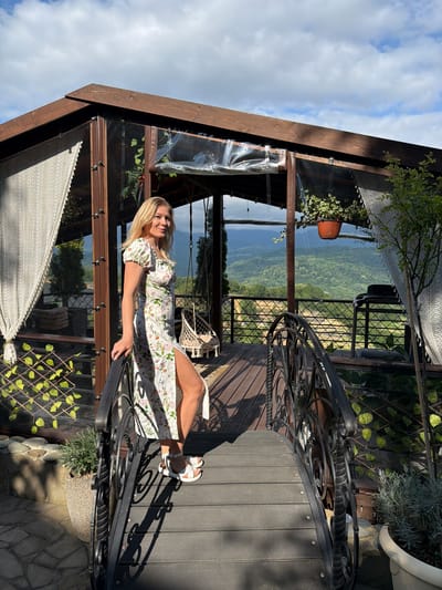 In Nizhnyaya Shilovka, Russia, a blonde woman stands on a wooden bridge. She is wearing a white floral print dress with a thigh-high slit and white sandals. The bridge has ornate black metal railings and is set against a backdrop of a wooden structure with clear plastic sheeting and white lace curtains. Beyond the structure, a lush green valley is visible under a cloudy sky. A hanging chair and potted plants are also present in the scene.