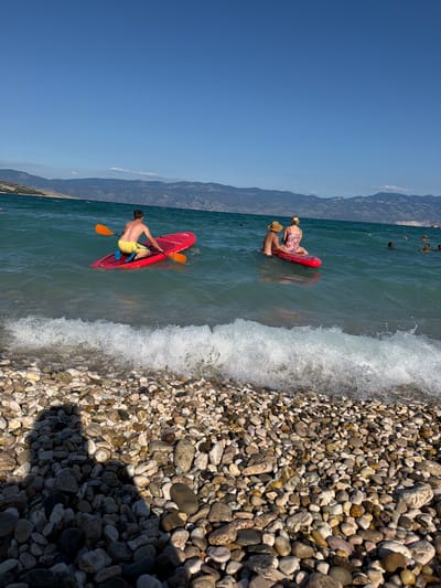 The image depicts a sunny day at a pebble beach in Baška, Općina Baška, Croatia. In the foreground, the beach is covered in smooth, multi-colored pebbles, with a gentle wave breaking at the waterline. In the middle ground, two individuals are on red stand-up paddleboards in the turquoise water. One person, wearing yellow shorts, is paddling a board with an orange paddle. Another person, wearing a patterned swimsuit and a straw hat, is sitting on a board with someone else behind them, also in the water. In the background, there are hazy mountains under a clear blue sky, suggesting a coastal mountain range. Several other people are visible swimming or floating in the water further out.