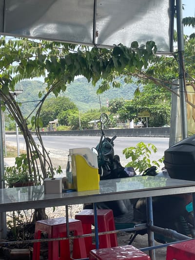 A low-angle perspective shows an outdoor setting in Nam Ninh Hoa, Vietnam. A road passes outside of a sheltered area, possibly a cafe or eatery, evident by the arrangement of red plastic chairs. A motorcycle with a top case and a yellow storage unit is parked, suggesting recent or intended usage. Foliage is present in the foreground and background, integrating built and natural elements. A sign is visible across the road.