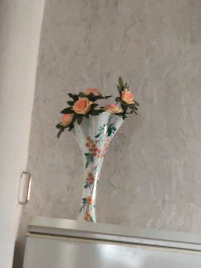 A vase of artificial flowers is displayed on a shelf against a textured wall. The vase is white with a decorative pattern of orange berries and green leaves. The flowers are coral-colored roses with green leaves. The shelf is silver and appears to be part of a larger appliance, possibly a refrigerator. The wall has a light grey textured finish. A silver handle is visible on the left side of the frame, attached to what looks like a door or cabinet. The scene appears to be indoors, likely in a residential setting. The location is not specified as being in the city of Karaganda, Russia.