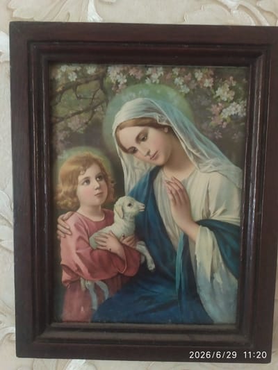 A framed painting depicts the Virgin Mary and the infant Jesus, accompanied by a lamb, in a natural setting with blossoming trees. The Virgin Mary, draped in a blue robe over a white garment and a light blue veil, cradles a young lamb with her left arm. Her right hand is raised, touching her chest. The infant Jesus, to the left, is shown in profile, wearing a pink tunic and gazing upwards. He has his left arm around the lamb. Both figures have a faint halo. The background features a soft, out-of-focus arrangement of tree branches with small pink and white blossoms against a muted green and yellow light. A dark wooden frame surrounds the painting. In the bottom right corner of the frame, a timestamp reads "2026/6/29 11:20". The painting is hanging on a textured wallpapered wall with a floral pattern. The image is captured from a slightly elevated angle.