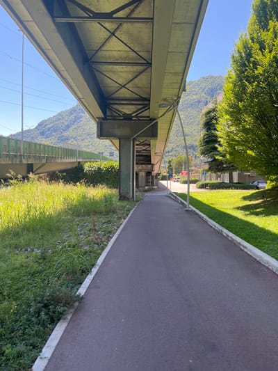 This is a landscape photograph taken from below a bridge or overpass, looking out towards a mountain and a paved path. The image is oriented vertically, with the sky and mountains appearing at the top and bottom respectively.
The main subjects are the structural elements of the bridge, a paved path, lush green grass and trees, and a distant mountainous landscape. The bridge's underside is visible on the left, showing metal beams and supports. To the right of the bridge is a dark gray paved path with a concrete border, bordered by a strip of green grass and wild vegetation. In the distance, a verdant mountain range rises against a clear blue sky. A section of a green-railed walkway or bridge is also visible further down, parallel to the main path and the mountain.
There are no people or distinct activities clearly depicted, suggesting a peaceful, possibly recreational setting. The lighting indicates it is daytime, with bright sunlight casting shadows on the grass and highlighting the foliage. The clear sky suggests fair weather.
The provided crop hints suggest a location context of Lecco, Italy, which aligns with the mountainous terrain visible in the background. There is some faint green graffiti on one of the bridge supports, but no other significant text is legible. The overall mood is serene and natural, with a focus on the interplay of man-made structures and the surrounding environment.