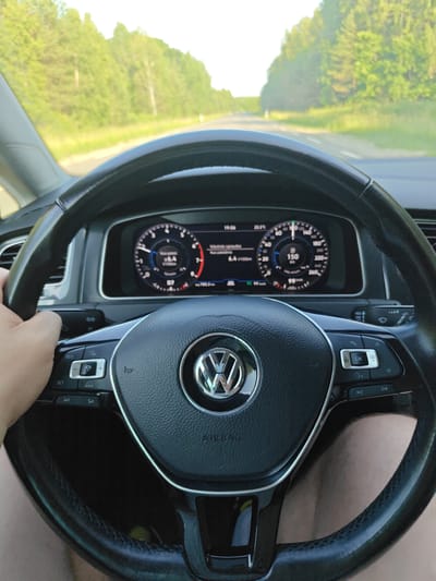 This is a first-person perspective image taken from inside a Volkswagen car, likely a Golf, as it drives on a rural road. The driver's left hand is visible on the steering wheel, which displays the Volkswagen logo and various controls. The dashboard features a digital display showing the time as 19:06, outside temperature as 25.5°C, current speed at 150 km/h, and average fuel consumption at 6.4 L/100km. The road ahead is flanked by lush green trees, suggesting a summer day in a scenic, possibly wooded area. Based on the context provided, this scene is located on a road near Traksėdžiai, Lithuania.