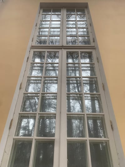 The image depicts two adjacent, multi-paned windows in a cream-colored wall. The windows are white-framed and feature a grid of small glass panes. Through the windows, bare trees are visible, their branches intertwining against an overcast sky. Hinges are visible on the tops and bottoms of the windows, indicating they are likely casement windows. The location is suggested to be Arkhangel'skoe, Russia, though this cannot be confirmed from the image alone. No people or actions are visible in the image. The environment appears to be outdoors, with a focus on the trees and the exterior of a building.