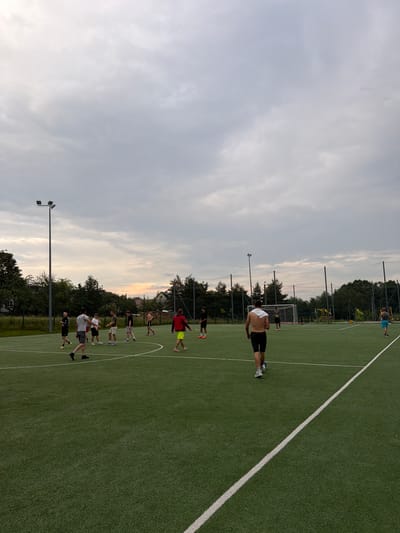 The image depicts a group of young men playing soccer on an artificial turf field. The field is marked with white lines, and a soccer goal is visible in the background. Several floodlights are positioned around the field, suggesting it can be used at night. The sky is overcast, with a hint of sunset visible in the distance, indicating it is likely late afternoon or early evening. The surrounding area is a mix of trees and residential buildings, which could be consistent with the outskirts of a town like Mordarka, Poland. The men are dressed casually in athletic wear, with some shirtless and others wearing t-shirts and shorts. Their actions suggest they are engaged in a friendly game of soccer, with some actively playing and others observing or preparing to join. The overall atmosphere appears relaxed and informal, typical of a recreational sports activity.