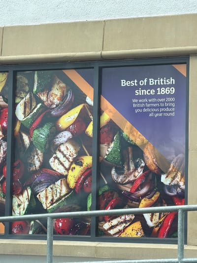 The image shows a poster or advertisement mounted on a building facade. The text on the poster reads "Best of British since 1869. We work with over 2000 British farmers to bring you delicious produce all year round." The poster also features a photograph of grilled vegetables. A metal structure, likely a ladder or scaffolding, is visible in the foreground, partially obstructing the view of the poster. The building's exterior is made of beige stonework or concrete. It could be a sign outside a store or restaurant that emphasizes the use of local products. The image was taken in Durham, United Kingdom.