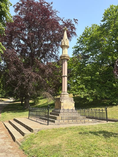 The image shows a stone monument or memorial cross situated in a green space, possibly a park or cemetery, in Ipswich, United Kingdom. The monument consists of a cylindrical column made of what appears to be pink granite or similar stone, topped by an ornate, lighter-colored stone capital and spire. The monument is surrounded by a low, black metal fence or railing. To the left of the monument is a large tree with deep red or purple foliage. Behind and around the monument and the red-leaved tree are other green trees and grassy areas. There are stone steps leading up to the monument's base. The sky is visible in the upper left corner and is a clear blue. The photo is taken during the daytime. No people are visible in the image.