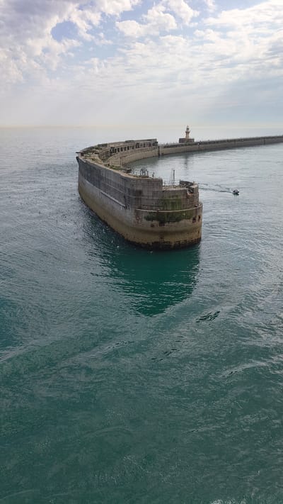 This is an aerial, vertical, wide-angle shot of a large, abandoned ship partially submerged in the sea. The ship is the SS Kampala, a former ferry that ran aground in Dover, United Kingdom. It is mostly dilapidated, with visible rust and decay. The ship is positioned diagonally across the frame, with its bow pointing towards the left and its stern towards the right. The sea is a deep turquoise color, with ripples and waves indicating a calm day.
In the background, a long, concrete pier extends into the distance. A small, white lighthouse is visible on the pier. There are also some buildings and structures along the pier, suggesting it is part of a harbor or port. On the left side of the frame, a cloudy sky dominates the upper portion of the image. The clouds are white and fluffy, set against a pale blue sky, creating a bright and somewhat hazy atmosphere. The overall impression is one of abandonment and decay against the backdrop of a calm, natural setting.
There are no people clearly visible in the image, though a very small boat can be seen in the water near the stern of the ship, and some seagulls are scattered around the ship. The time of day appears to be daytime, likely morning or afternoon, given the brightness of the sky and the clear visibility. The weather appears to be fair with light winds. There is no visible text in the image.