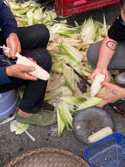 Two individuals are in the process of shucking corn, with husks and silks scattered on the ground. One person, wearing dark pants and a dark long-sleeved shirt, holds a partially shucked ear of white corn in their left hand while using their right hand to peel away the remaining husk. They are wearing green canvas shoes. The other individual, wearing grey pants and a black t-shirt, has a circular tattoo with a dot in the center on their left forearm and a doodle tattoo on their right hand, which is holding a shucked ear of white corn. A red bucket or container is partially visible in the upper right background, and a woven basket and a blue plastic basket are in the foreground. The scene appears to be outdoors, possibly in a market or roadside setting in Chongzhou City, China, given the context.