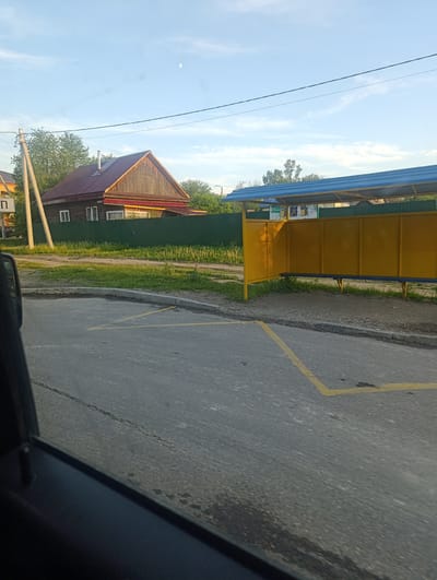 The image depicts a rural street scene in Svobodny, Russia, during daylight. The foreground is dominated by an asphalt road with yellow markings that suggest a parking or waiting area. On the right side of the road, a yellow bus stop shelter with a blue roof provides a place for people to wait. A blue bench is visible inside the shelter. Behind the bus stop, a green fence encloses a grassy area and trees.

In the background, to the left of the bus stop, a traditional wooden house with a red roof stands. The house has several windows and appears to be a residential dwelling. A utility pole with wires is situated in front of the house, and a sign with a Cyrillic letter "П" is attached to it. The sky is partly cloudy, with a sliver of moon visible. The overall impression is one of a quiet, everyday scene in a Russian village or small town. The lighting suggests it is daytime, possibly late afternoon or early morning, given the gentle sunlight. No people are visible in the frame. The weather appears fair.