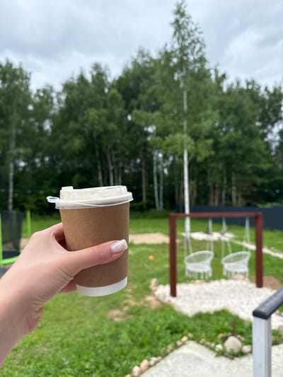 A hand holds a disposable brown paper cup of coffee in front of a blurred outdoor scene in the city of Ryzhkovo, Russia. The background features a line of birch trees, a playground with two white wicker swings hanging from a wooden frame, and green grass. The sky is overcast.