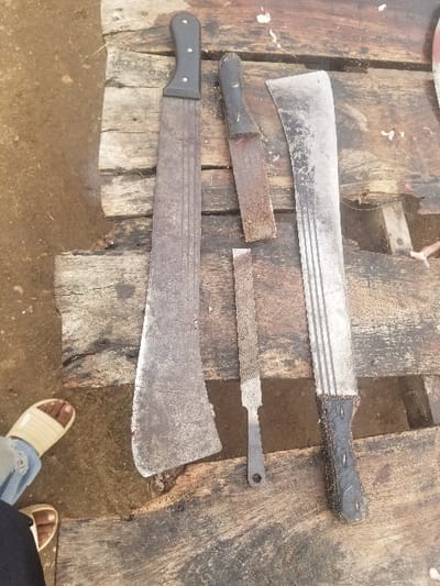 The image shows three large knives and a metal file arranged on a wooden surface, likely a table or counter. The knives are all large and appear to be made of metal, with wooden handles. The file is also made of metal, and has a wooden handle. The knives are arranged vertically on the surface, with the file placed between them. The surface is made of rough wood planks that are worn and stained. The environment is likely outdoors.  The image does not include any people.