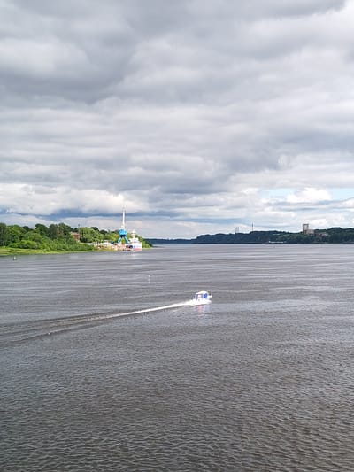 A body of water stretches from the foreground to the horizon under a cloudy sky. A motorboat with a white hull and a blue-trimmed cabin is moving across the water, leaving a white wake behind it. The left bank is covered with dense green foliage. A crane stands on the bank with a white tower and blue supports, along with a cargo ship, likely at a port facility. In the distance on the opposite bank, there are some buildings and transmission towers. The image is likely taken from a high vantage point, like a bridge or elevated riverbank. The location is the city of Rybinsk, Russia.