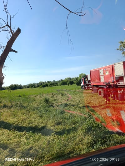 The image depicts a rural scene in Danceni, Moldova, on June 17, 2026, at 09:58. A large red fire truck, equipped with a generator unit, is parked in a grassy field. Several firefighters, dressed in dark uniforms with yellow accents, are standing near the truck, seemingly engaged in an operation. Red hoses are laid out across the field, extending from the truck. The environment is characterized by a clear blue sky, lush green vegetation, and a few trees in the background. A dead tree with broken branches is visible on the left side of the frame. The overall impression is one of a coordinated response to an incident in an outdoor setting.