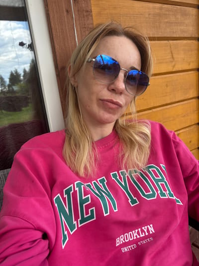 A woman with blonde hair and sunglasses is photographed outdoors, sitting in front of a wooden wall. The woman is wearing a bright pink sweatshirt with "NEW YORK" and "BROOKLYN UNITED STATES" printed on it in green and white letters. The background shows a window with a reflection of trees and sky. This image was taken in Shuiskaya Chupa, Russia.