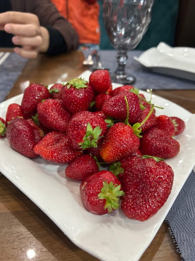 A close-up shot of a square white plate filled with fresh strawberries. The strawberries vary in size, with some having green stems attached. The plate is placed on a wooden surface with a blue and white patterned cloth visible. A hand is partly visible in the lower left corner, reaching towards the plate. A glass object, possibly a vase, is blurred in the background on the left side of the image. The photo does not provide information about the city of Ақсу, Kazakhstan.