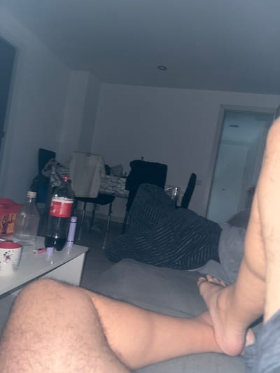 The image captures an indoor scene, likely a living room or bedroom, from a low perspective, rotated 90 degrees counter-clockwise. The lighting is dim, suggesting it is evening or night.
In the foreground, a person's bare legs and feet are visible, resting casually, implying a state of relaxation. One foot is positioned on top of the other leg, and the skin has a light complexion with visible hair. The person appears to be lying on a grey sofa or bed, parts of which are visible on the right side of the frame.
In the mid-ground and background, various household items are arranged. A white table or shelf holds several objects, including a large, dark plastic bottle of Coca-Cola with a prominent red label, and another clear plastic bottle, possibly empty. Text visible on the Coca-Cola label includes "RECIKLAM". Also on the table are two purple/pink cylindrical objects (possibly vape pens or similar devices) and a white mug with a red handle. A dark, striped blanket or fabric is draped over a chair or sofa in the mid-left. Further in the background, a black office chair is partially visible, and beyond that, a window or glass-paneled door can be seen, offering a glimpse of what appears to be an exterior or another room. The walls are a light, neutral color, with a small black circular object (possibly an outlet or sensor) on one wall.
The scene suggests a casual, relaxed atmosphere in a residential setting in Móstoles, Spain.