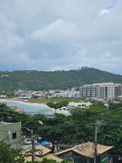 An elevated long shot captures a coastal scene in Arraial do Cabo, Brazil, under an overcast sky. The foreground features a utility pole with multiple electrical wires and insulators, positioned above the tiled roofs of several low-rise structures and obscured by dense green tree foliage. To the lower left, a portion of a two-story green building with a dark rectangular window and a circular white emblem is visible.
The midground reveals a light blue-green bay with a curved, sandy white beach. Multiple small boats, some with orange hulls, are anchored or moving in the calm water. A pier extends into the bay, with several individuals visible on its structure and in the shallow water near the shore. The beach itself shows numerous colorful beach umbrellas (red, orange, blue, green) and scattered beachgoers, indicating active recreational use.
Behind the beach area, a cluster of residential and commercial buildings forms a coastal town. These structures vary in size and architectural style, including a prominent multi-story building with a beige facade and numerous balconies on the right side of the bay. Roads or pathways are visible between these buildings and the beach.
The background is dominated by a large, verdant hill covered in dense green vegetation and scattered with several small buildings and houses along its slopes and crest. The sky above is entirely covered with diffuse grey and white clouds. A small dark speck, potentially an avian subject, is visible in the upper left portion of the sky.