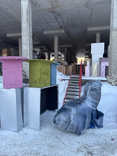 The image displays a winter scene in L'vovskiy, Russia, situated on snow-covered ground adjacent to or beneath an unfinished concrete structure. The structure features exposed concrete pillars and a flat, unpainted concrete ceiling.
In the foreground, from left to right, three square-topped pedestals or tables, upholstered in velvet-like fabric, are positioned on top of silver-colored, rectangular box-like units. The pedestals are pink, lime green, and light blue. One silver box beneath the blue pedestal is open, revealing a dark interior. To the right, a dark blue armchair, completely encased in transparent plastic shrink wrap, rests directly on the snow.
In the mid-ground, a temporary staircase with red metal frames and wooden steps descends into the concrete structure; snow is visible on and around the steps.
The background, within the confines of the concrete structure, contains various materials. To the left, wooden pallets and other construction supplies are stacked. To the right, a stack of rectangular boxes, some white, others covered in pink or gold glitter, are arranged against a concrete pillar. Distant, indistinct shapes or figures are visible deeper within the structure, illuminated by ambient light from an unseen opening. No immediate human activity is observed in the primary view.