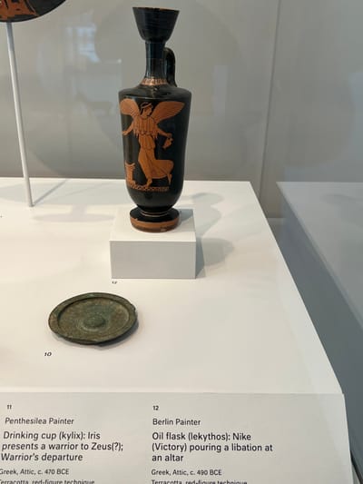 The image displays a museum exhibit at the Harvard Art Museums in Cambridge, Massachusetts. The central focus is an ancient Greek Attic terracotta oil flask (lekythos) dated approximately 490 BCE, attributed to the Berlin Painter. This black-ground vessel, featuring a red-figure technique, depicts a winged figure identified as Nike (Victory) pouring a libation at an altar. The lekythos rests on a small, white rectangular pedestal situated on a larger white display surface. Below and to the left of this pedestal, a circular, patinated bronze object with a central raised boss, possibly a mirror or dish, lies directly on the white surface. At the upper left, the curved rim of another ancient Greek artifact, identified by an adjacent label as a red-figure terracotta drinking cup (kylix) by the Penthesilea Painter (c. 470 BCE), is partially visible. The background consists of plain light gray walls and white display structures, with a thin white vertical support rod visible on the left. Item numbers "10", "11", and "12" are printed on the lower white display surface, identifying the artifacts.