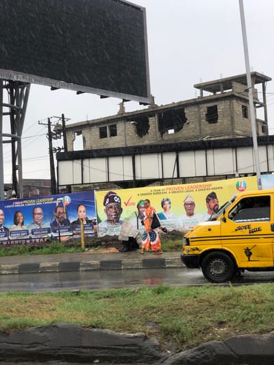 The image depicts a street scene in Alagbede, Nigeria, characterized by heavy rainfall, with visible raindrops against the lens. In the foreground, a yellow commercial van, identified by the text "SOTE NILE" on its side, is moving to the right, its wheels splashing water on the wet asphalt road. Two women are walking on the sidewalk to the left of the van, one in an orange and white patterned dress and the other in a more neutral attire.

Behind the sidewalk, a row of large political banners is displayed. The banners feature portraits of various individuals, some wearing traditional attire, and slogans such as "A TEAM OF PROVEN LEADERSHIP" and "A GREATER SURULERE, A GREATER LAGOS, A GREATER NIGERIA." The "APC" logo is visible on one of the banners, indicating a political affiliation.

Further in the background, a partially constructed or derelict concrete building stands with exposed rebar and incomplete walls, its structure appearing damaged. Above and to the left of this building, a large billboard structure is present, with the text "INVENTMEDIA LIMITED" visible on its frame. The sky is overcast, consistent with the rainy weather. The overall environment suggests an urban setting undergoing development or experiencing economic hardship.