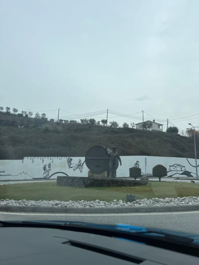 The image displays a roundabout in Santa Marta de Penaguião, Portugal, viewed from inside a vehicle. In the center of the roundabout, a sculpture depicts a male figure wearing a hat, standing alongside a large, dark-colored barrel positioned on a stone base. Behind this monument, a curved white wall features black line-art murals, illustrating grapevines, a person harvesting grapes, and the outline of a building and another figure. The central island of the roundabout is covered in green grass, bordered by a ring of light-colored rocks, and includes two dark, trimmed hedges to the right of the main sculpture.

A steep, sparsely vegetated hill dominates the background, with scattered small trees visible across its surface. A white house with a dark roof stands on the upper portion of the hill to the right. Utility poles and multiple power lines span horizontally across the mid-ground. The sky is overcast and uniformly light grey. The bottom foreground shows the top edge of a car's dashboard and windshield, with a blue reflection or object visible on the dash.