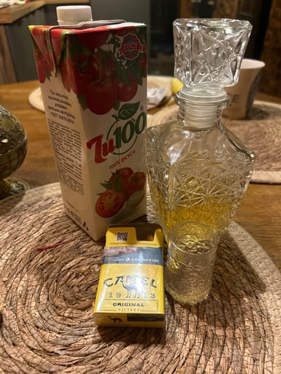 The image depicts a still life arrangement on a woven placemat, likely on a wooden table. The scene includes a carton of tomato juice, a crystal decanter filled with a golden liquid and a small glass containing the same liquid, and a pack of Camel cigarettes. The tomato juice carton is red and white with images of tomatoes and text in a Cyrillic script. The decanter is ornate with a faceted stopper and a diamond-patterned body. The glass is short and also features a faceted design. The Camel cigarette pack is yellow and displays the brand's logo. In the background, a portion of what appears to be a globe or decorative sphere is visible, along with a mug and the edge of a table. There are no people or animals in the image. The lighting suggests an indoor environment. The text on the cigarette pack mentions "НАВЕДИ КАМЕРУ, ЧТОБЫ УЗНА..." which translates to "POINT THE CAMERA TO LEARN...". There is no indication of the city of Staryy Otrada, Russia.