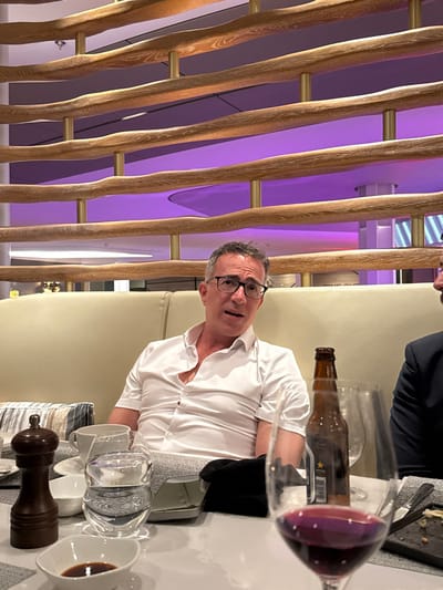 An indoor dining scene features two individuals seated at a white table in what appears to be a contemporary restaurant or lounge in Rum Cay, Bahamas.
The central figure is a male, estimated to be in his 50s or 60s, with short, curly grey hair and black-framed eyeglasses. He is wearing a white short-sleeved collared shirt, with the top buttons unfastened, and is seated in a light beige upholstered booth. He faces forward, looking towards the camera, with his mouth slightly ajar, suggesting he is speaking or about to speak. To his immediate right, a second individual's left shoulder and a portion of their dark blue or black suit jacket are visible, indicating another person is seated at the table.
The white tabletop is populated with various dining items: a dark wooden pepper mill, a small white rectangular dish containing a dark liquid (possibly soy sauce), a striped blue and white cloth napkin, a white ceramic cup, a clear glass filled with water, and a smaller white bowl. Towards the right, a metallic-silver-colored clutch or purse rests next to a folded black napkin. A brown glass beer bottle, bearing a white label (partially obscured, but the brand "Sapporo" is faintly discernible), stands upright. In the immediate foreground, a wine glass filled with red wine occupies the right side of the frame, slightly out of focus. A plate with minimal food residue is also visible on the far right.
The background consists of a decorative architectural element: a partition or wall feature composed of multiple horizontal, undulating wooden planks or branches, supported by vertical brass-colored rods. Behind this structure, a smooth, curved surface is illuminated with a consistent purple ambient light, suggesting a modern interior design.