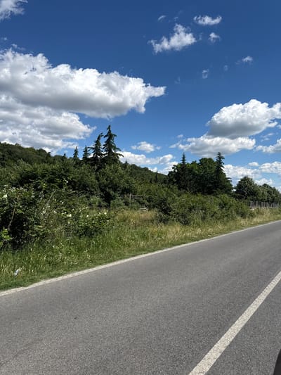 The image shows a paved road in the city of Sotirya, Bulgaria, on a bright, sunny day. The road is empty, with a single white line marking the lane. To the left of the road is a grassy embankment covered in lush green vegetation, including bushes and trees. In the background, a densely wooded hill rises, topped with a bright blue sky filled with scattered white clouds. The overall scene is serene and natural, suggesting a quiet, rural setting.