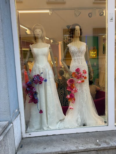 The image shows a storefront window display featuring two mannequins dressed in wedding gowns. The mannequins are positioned inside a brightly lit interior, with the exterior environment reflected in the glass. The left mannequin wears a strapless white gown with a textured fabric and a full skirt, adorned with a cascading arrangement of purple and blue flowers. The right mannequin is adorned with a lace wedding gown featuring a plunging neckline and a train, decorated with a vibrant bouquet of pink and orange flowers. Behind the mannequins, several people are visible, including a woman in a black and white striped dress and another person seated on a red sofa. A neon sign with the words "Welcome to the Jungle" is visible in the background. The storefront appears to be located in Würzburg, Germany, as indicated by the presence of a sign that says "Würzburg" in the lower right corner of the image, partially obscured by the window frame. The exterior of the building features a blue-gray textured wall and a concrete sidewalk in the foreground.