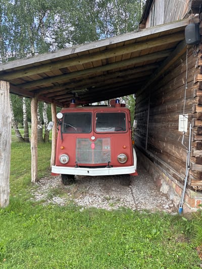 A red vintage fire truck is parked under a wooden shelter next to a rustic wooden building. The truck is positioned facing forward, with its headlights, grille, and red emergency lights visible. The shelter's roof is constructed of wooden planks, supported by rough-hewn wooden posts. To the right of the truck, the wooden building features weathered planks and a window. A white sign with text is affixed to the building's exterior. The ground in front of the truck is covered with gravel and stones, while the surrounding area is overgrown with green grass. In the background, trees with green foliage are visible. The scene appears to be located in Sielachowskie, Poland.