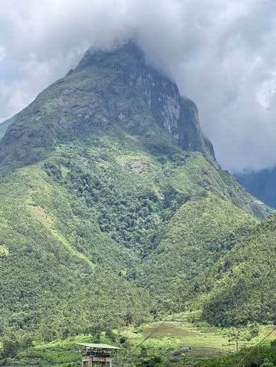 The image captures a dramatic mountainous landscape near the city of Bình Lư, Vietnam. The foreground features a man-made structure, possibly an observation deck or a small building, elevated on concrete stilts. This structure is situated on a verdant, gently sloping hill. Beyond this, the terrain rises sharply, covered in dense, lush green vegetation that blankets the steep mountainsides. The upper reaches of the mountains are partially obscured by thick, grey clouds, lending a dramatic and somewhat mysterious atmosphere to the scene. The overall impression is one of untamed natural beauty, with a touch of human intervention visible in the foreground.