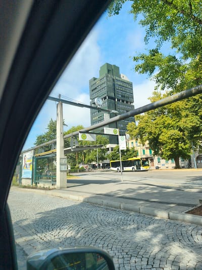 The image depicts a street scene in Lörrach, Germany, as viewed from inside a vehicle. In the foreground, a cobblestone street leads towards a bus stop. A modern, dark-colored building with many windows and a green top section dominates the background. A large clock face is visible on the side of this building. To the right of the bus stop, a yellow and white bus is parked. Trees with green foliage are present on both sides of the street. The sky is blue with some scattered clouds. Several people are visible at the bus stop, including one individual sitting on a bench inside the shelter. The overall impression is of a typical urban environment on a sunny day.