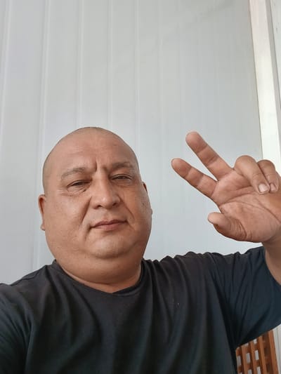 A man with a shaved head, wearing a black t-shirt, is captured in a selfie. He is making a peace sign with his right hand, with two fingers extended. His eyes are partially closed, and he has a slight smile on his face, suggesting a relaxed or contented mood. The background is a simple, light-colored wall with horizontal lines, possibly indicating blinds or a similar textured surface. The lighting suggests it is daytime, and the indoor setting is implied by the smooth, even background. The composition is a close-up, focusing on the man's face and hand. No text is visible in the image. The photo appears to have been taken in Gulistan, Uzbekistan.