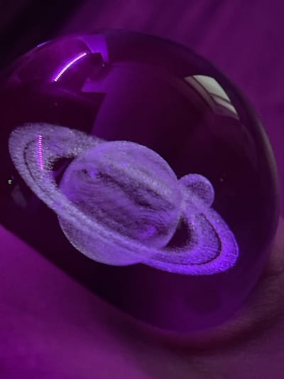 The image displays a close-up, slightly out-of-focus view of a glass orb containing a 3D etched model of the planet Saturn, illuminated by purple light. The orb is held by a person whose hand and arm are visible in the background, out of focus and also bathed in the purple glow. The texture of the orb is smooth and reflective, with highlights from the lighting. The etched Saturn model is detailed, showing the planet's rings and its spherical body. Small, faint green lights are visible near the bottom of the orb. The overall mood is somewhat ethereal and mysterious due to the dominant purple lighting and the celestial subject matter. There is no visible text in the image. The context provided indicates the location is Gliwice, Poland, but this cannot be inferred from the visual content of the image itself. The time of day and weather are not discernible.