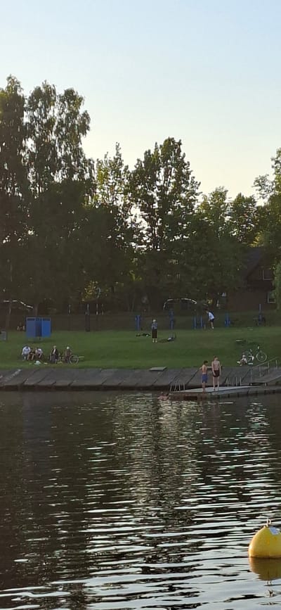 The image depicts a waterfront scene, likely in Pärnu, Estonia. Several people are present, some standing on a concrete dock, while others are on a grassy area next to it. The water is calm, reflecting the sunlight. On the grass, there are several bicycles, suggesting a recreational or leisure activity. A yellow buoy is visible in the water in the upper right corner of the image. The time of day appears to be late afternoon or early evening, as indicated by the quality of light. The presence of people in swimwear and the overall atmosphere suggest a warm weather day at a public swimming area or recreational spot. The background features lush green trees and a clear sky, contributing to a peaceful, summery ambiance. There is no visible text in the image.