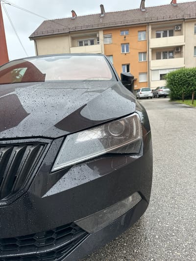The image depicts the front right quarter of a dark-colored vehicle, likely a sedan or hatchback, parked on an asphalt surface in a residential area of Maribor, Slovenia. The car's surface, including the hood, headlight, grille, and bumper, is covered in numerous small water droplets, indicating recent rain. The prominent right headlight, a black multi-slat grille, and a lower hexagonal mesh intake are visible. A small rectangular sensor is present on the bumper below the headlight.
In the background, a two-story beige building with a brown tiled roof and a red chimney is situated to the left. To the right of that, a multi-story residential building features an orange facade on its left section and a lighter beige facade on its right. This building has multiple windows, some with external blinds, and several balconies. Two air conditioning units are mounted on the beige section of the building. Further back, two light-colored cars are parked on the wet asphalt, one appearing to be a white hatchback and the other a silver sedan. A dense green hedge borders the right side of the scene, separating the paved area from a grassy patch. The sky is overcast, and no individuals are present in the frame.