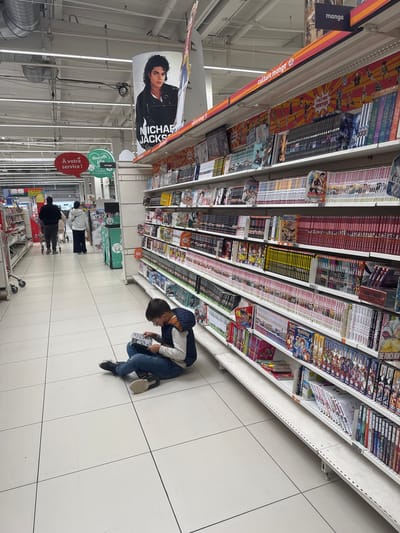 The image depicts the interior of a supermarket in Leers, France, with a prominent display of manga and anime merchandise. In the foreground, a young boy wearing glasses, a blue vest, and jeans is sitting on the tiled floor, engrossed in reading a comic book. He is positioned to the right of the frame, amidst a vast selection of manga volumes displayed on white shelving units. The shelves are filled with numerous books, some featuring colorful covers with anime characters. Above the manga section, signage reads "manga" and "culture manga." Further down the aisle, two individuals, a man and a woman, are seen walking away from the camera, pushing a shopping cart. In the background, the supermarket's ceiling with industrial lighting and hanging displays is visible. One of these displays is a large poster of Michael Jackson. Adjacent to this, speech bubble-shaped signs read "À votre service !" and "Nouvelle Vie manga." The overall environment suggests a retail space dedicated to entertainment and lifestyle products.