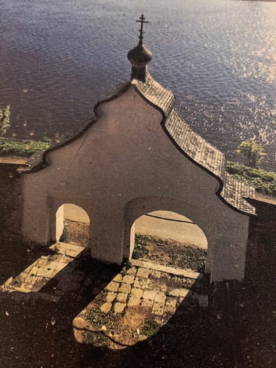 This is an eye-level medium shot of an archway structure on the shore of a body of water, likely a river, in Staraya Ladoga, Russia. The structure has a light-colored, textured facade with two arched openings. Above the openings, the roof is tiled and curves upwards to a central, onion-domed spire topped with a cross. Sunlight casts strong, defined shadows of the arches and the structure onto the cobblestone ground in front of it, where patches of grass are visible. The body of water behind the structure is dark blue with sunlight reflecting off its surface, creating shimmering patterns. The shoreline is lined with greenery and some scattered trees. The image appears to be a photograph, possibly old, given its texture and slight graininess.