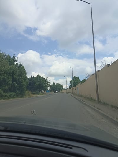 The image captures a view from inside a vehicle, looking out onto a road. The scene appears to be set in Red Sloboda, Azerbaijan. The foreground shows the dashboard and the Mercedes-Benz emblem on the hood of the car. The road ahead curves to the left, with a concrete barrier on the right side and trees and vegetation on the left. A long, tan-colored wall runs along the right side of the road, with a lamppost in front of it. The sky is partly cloudy with large white cumulus clouds against a blue backdrop. Several other lampposts are visible along the road, and a few vehicles are in the distance. The overall impression is that of a drive through a suburban or semi-rural area.