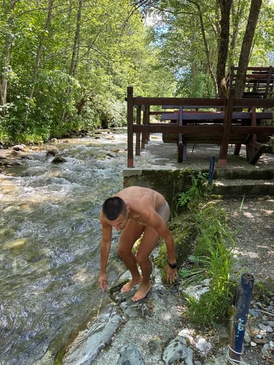 In this vertical, medium-shot photograph taken at Bare Kraljske, Montenegro, a shirtless man wearing light-colored shorts is partially submerged in a shallow, fast-flowing river. He is crouching on a rocky bank, with his upper body leaning forward and his hands extended into the water. His gaze is directed downwards at his hands, as if he is feeling the cool water or collecting some. His skin appears tanned, and he has short dark hair. His feet are visible, also wet and positioned on the rocks.

The river is clear and rocky, with ripples and small waves indicating movement. The surrounding environment is lush and green, with trees and foliage densely covering the banks. Sunlight filters through the leaves, creating dappled patterns on the water and the ground. In the background, to the left, is a wooden structure, possibly a picnic table or seating area, with dark wooden planks. Concrete steps lead down towards the river from this structure. The overall atmosphere suggests a warm, sunny day in a natural, serene setting. The man’s posture and focus on the water might indicate a moment of cooling off or enjoyment of the natural elements. No visible text is present in the image.