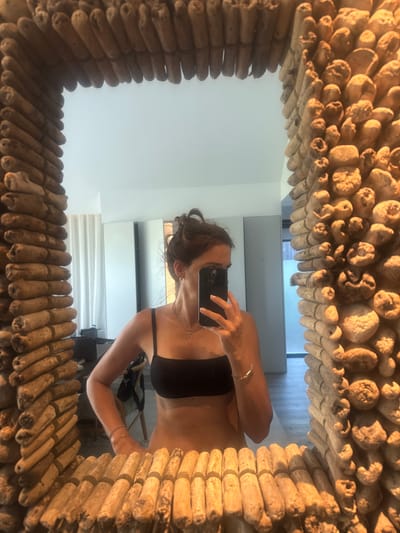 A woman is taking a mirror selfie in a room that appears to be a hotel. The mirror has a thick, rustic frame made of stacked wooden pieces. The woman is wearing a black bra and has her hair tied up. She is holding a smartphone in her right hand to take the picture. The background shows a brightly lit room with white curtains and some furniture, possibly a desk and a chair. The flooring is a light color. There are no specific landmarks visible that would definitively place this image in Grândola, Portugal.