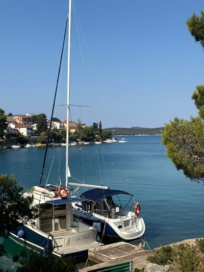 The image depicts a serene harbor scene in the city of Šibenik, Croatia, on a clear, sunny day. The foreground is dominated by two docked boats: a smaller white and blue vessel with the name "Sv. Roko" visible on its stern, and a larger sailboat with a prominent mast. Both boats are moored in calm, turquoise water. In the background, a picturesque coastline is lined with houses, their red-tiled roofs contrasting with the lush green trees and vegetation. The distant horizon is marked by rolling hills under a vast blue sky. Several other smaller boats are scattered across the water, and a few people can be seen swimming or kayaking in the distance, enjoying the tranquil aquatic environment. The overall impression is one of peaceful seaside living and recreation.