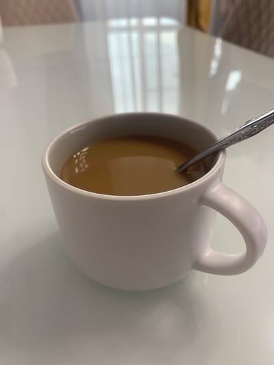 A white ceramic mug, filled with a light brown liquid, rests on a white, reflective surface. A metal spoon, with intricate scrollwork on its handle, is partially submerged in the liquid within the mug. The background is blurred, showing hints of vertical blinds and possibly furniture. The scene is an indoor setting.